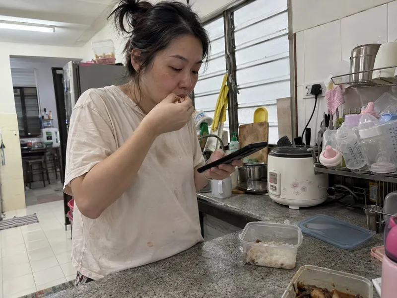 产后妈妈下午站在厨房，一手吃剩饭一手看手机，旁边晾着奶瓶